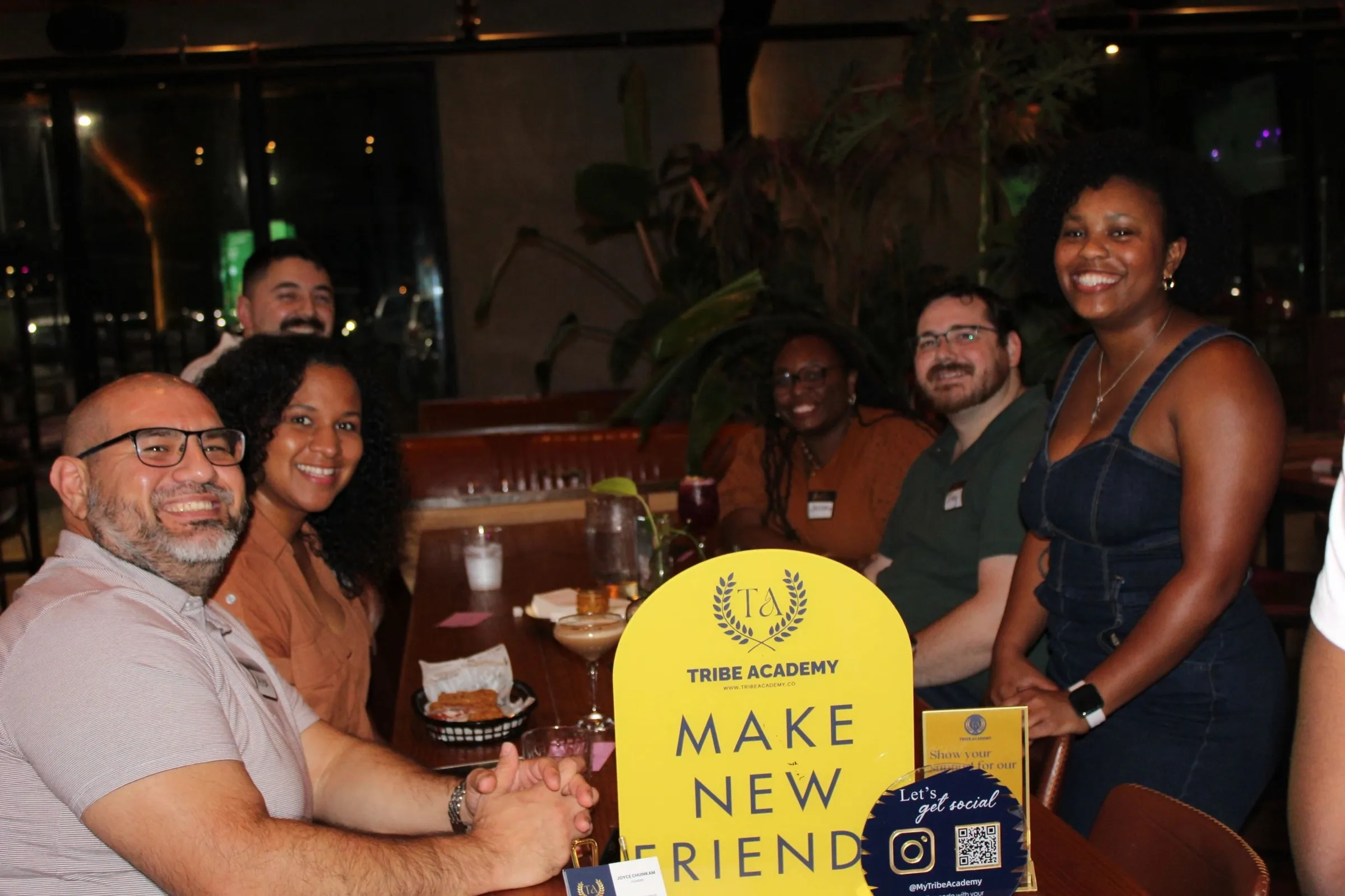 Members laughing around a table beside a yellow Tribe Academy "Make New Friends" sign.